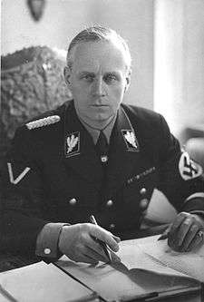 A blonde-haired male wearing a dark Nazi Party uniform with swastika armband seated at a desk