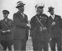 Five men, two in the foreground and three in the background, one mostly obscured. Two men are in suits and three are in their military dress uniforms. All of the men in the picture are wearing hats. The two men in the foreground are shaking hands.