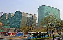 Two irregularly shaped greenish glass-faced buildings rise over a street lined with freshly budding trees on which a blue and white trolleybus is stopped