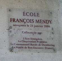 A clear plaque on a grey wall stating "École François Mendy" at the top with smaller writing below