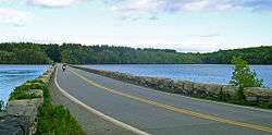 Ground-level view of a causeway crossing a lake. On either side of the causeway are short stone walls. The lake's banks are surrounded by thick woods.