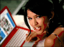 Image shows a bed with a switched-on red laptop. Beside the laptop lies a dark-haired woman in a sleeveless top, wearing large circular earrings while her hair is tied up with a fringe covering the left side of her face.