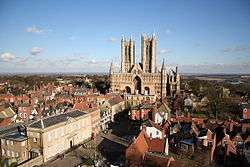 Aviw of Castle square - which is not a square at all. In the foreground is the cobbled surface of the open space which is surrounded by historic buildings. From left to right, the Regency Assembly Rooms, a Georgian three-storey house, Leigh-Pemberton house: a half-timbered building with the upper storey projecting, a medieval church, and the entrance to the Cathedral Close. In the left distance the towers of the Cathedral rise into a blue sky.