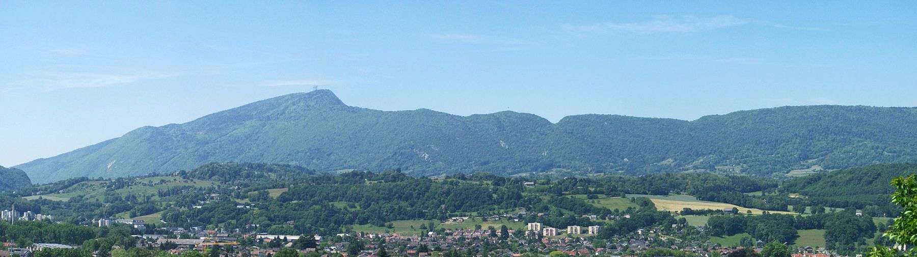 A detailed panorama of a long, notched, wooded ridge with a prominent peak toward its left-hand (southern) end. Fields occupy the lower hills in the middle ground, with a small town in the foreground.