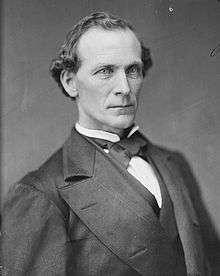 A man with curly, receding black hair wearing a black jacket, vest, and bowtie with a white shirt