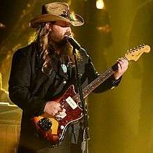 A blonde man with a hat is playing a guitar while singing on a microphone