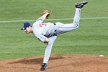 A man in a grey baseball uniform with a blue cap throws a baseball.