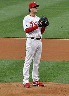 A man in a white baseball uniform with red trim. He is wearing a red cap and holding his left hand behind a black baseball glove.