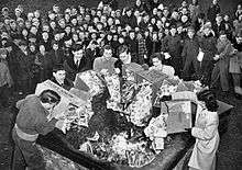 Photo of men and women burning boxes of comics in a bin as a crowd of children watch.