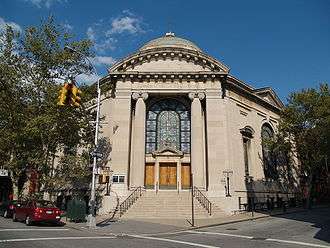 The front entrance of a hexagonal building capped by a dome is visible, facing a street-corner. The entranceway is framed by large stone columns and flanked by metal seven branched menorahs on each side. There are four wooden doors, one on each side and two in the middle, topped by a large arched stained-glass window. A stone stairway with metal railings on each side leads up from the sidewalk to the doors.