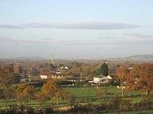 Roofs of houses showing amongst tree with prominent church tower. In the foreground are green fields with hills behind.