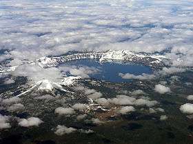 An aerial view of a circular lake ringed by continuous rock walls. Fleecy clouds partly obscure the view of the lake and the land beyond.