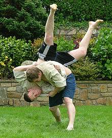 Two men in combat appear on a green lawn in front of a  shrubbery. Both men are wearing dark coloured shorts and cream coloured tops. One man has a tactical advantage, and is throwing his opponent head-first towards the ground.