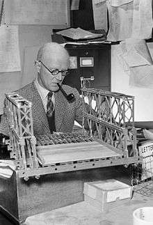 A middle-aged man sits at a desk while smoking a pipe; he is examining a model bridge in front of him