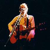 A male vocalist (wearing a cream shirt) singing into a microphone on stage while playing guitar