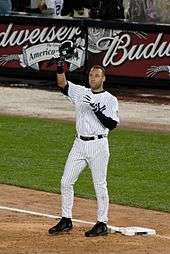 A man in a white baseball uniform with navy pinstripes removes his helmet to salute the crowd, which is cheering for him.