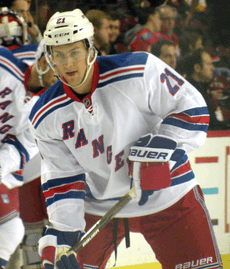 An ice hockey player leaning forward with his ice hockey stick resting on the ice. He is wearing a white helmet and a red, white and blue uniform.