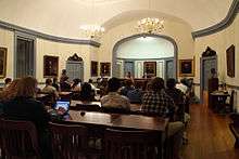 A collection of people sitting in a white room that has paintings on its walls.