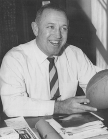 Dick Gallagher sitting at a table with a football in his hands in 1959