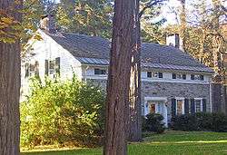 A stone house with white wooden sides and a narrow upper story also in white. There are two small chimneys at either end of the roof. Large tree trunks are in front; the house is lit with late-afternoon sunshine from the left