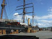 Partial view of a ship moored to a quayside. Prominent visible features are a mast with three crossbeams, two smaller masts, a funnel, a lifeboat and rigging. Packing cases are lined up on the quay, and a gangplank with "RRS Discovery" on it leads to the ship.