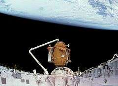 A space shuttle payload bay, covered in white insulation, with a small, cylindrical orange module at one end, supported by the shuttle's robotic arm. The blackness of space and the Earth serve as the backdrop.