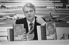 A black and white photograph of a middle-aged white man. He has grey hair and is wearing a black suit and tie. He is sitting at a table, and in front of him are two upright books; both  carries the face of Steve Biko, a young black man.