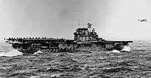 Black and white photo of a flat-decked ship at sea. It is carrying aircraft on the rear of its deck, and one plane is flying immediately in front of the ship.