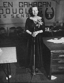 A well-dressed, tired looking woman stands before a microphone, part of a campaign banner "Helen Gahagan Douglas" is visible in the background.