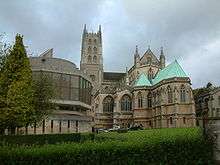 Ornate building with central tower. To the right is a stone building with green roof and to the left a new building with large glass windows.