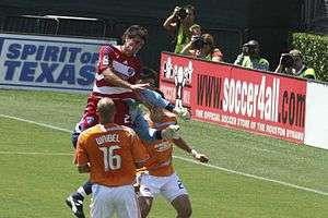 A FC Dallas player jumping in the air versus two Houston defenders and their goalkeeper