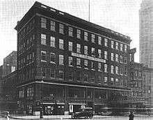 A six-story dark brick building for retail stores (street level) and offices (above), a hundred feet long and about 30 feet deep, behind which the theater wing stretches to the left. Between the fourth and fifth floors, a big sign on the facade says "Earl Carroll Theatre" in capital letters. The right side of the office building has a marquee, which reads "Just Because / A Melody Comedy", over the theater entrance, which is through the office building; there's another marquee at the theater wing on the left. An old-fashioned automobile is parked in front of the building.