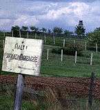 Behind a faded white sign saying "HALT ZONENGRENZE", there is a derelict barbed-wire fence supported by leaning wooden posts; some distance behind that, there are two newer-looking barbed-wire fences supported by concrete posts. Another fence and a guard tower are visible in the distance.