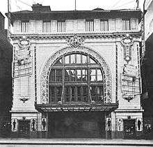 Black and white photo of the inside of a theater, with a high vaulted ceiling and an ornate curtain in front of the stage.