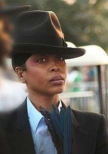 An image of a woman with a fedora and a suit holding blue feathers while staring forward with a neutral expression.