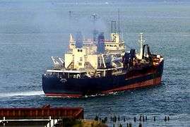 A large, almost rectangular black ship with a red hull creates a mild wake as it moves away from the shore into a very large river. Smoke rises above its decks, which are cluttered with antennas, mechanical fittings, and what appear to be self-contained rooms with multiple windows.