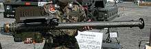 A large pipe-like weapon with a box on its lower lift hand side and upper left hand side, held up for display to the camera. A sign written in Japanese and a military truck can be seen in the background.