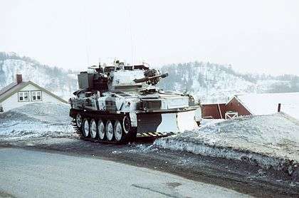 Vehicle in white winter camouflage. House roofs can just be seen behind and in the distance a range of snow-covered mountains