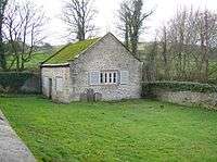 A small, simple chapel seen almost end-on in a grassy burial ground; it is built in stone with a moss-covered roof. On the end is a window with open shutters; on the front face is a door and two shuttered windows.