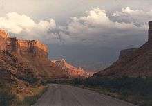 Highway through a sandstone gorge, with sunlight peering through clouds illuminating towers in the background.