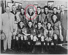 Two men in suits and another man in a Boy Scout uniform stand beside 10 seated teenaged boys in Boy Scout uniforms. Ford is indicated by a red circle.