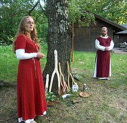 A man and a woman stand outdoors by a tree, wearing red and white robes.