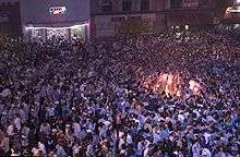 A large gathering of people on a street with a bonfire in the right side of the image.