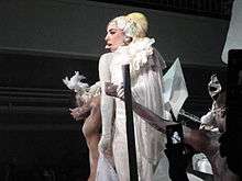 Gaga photographed from her lefft, wearing a white dress and flanked by her dancers.