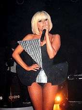 Gaga in a short dress with a triangular piece attached to her waist, singing into a microphone.