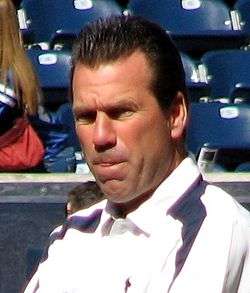 Color head-and-shoulders photograph of white man with dark (Gary Kubiak), wearing a white and navy sport shirt, squinting in bright sunlight, with stadium seating in the background.