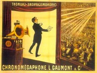 Illustration of a theater from the rear right of the stage. At the front of the stage a screen hangs down with the projected image of a tuxedoed man holding up a text and performing. In the foreground is a gramophone with two horns. In the background, a large audience is seated at orchestra level and on several balconies. The words "Chronomégaphone" and "Gaumont" appear at both the bottom of the illustration and, in reverse, at the top of the projection screen.