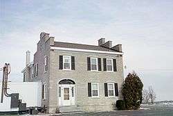 A two-story stone house with five windows and a door on its front side. Each window is flanked by shutters. A large tree is to the right of the house.