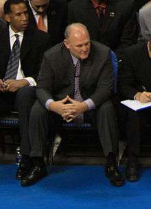 A man with gray hair, wearing a black suit, white shirt and tie, sitting at a basketball game.