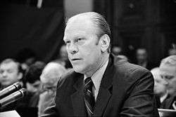 A man in a suit is seated at a table as he speaks into a bank of microphones. An audience is visible behind him.